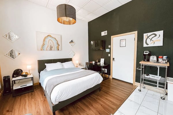 Modern bedroom with dark green accent wall, queen bed with white bedding, pendant light, wood flooring, and kitchenette area