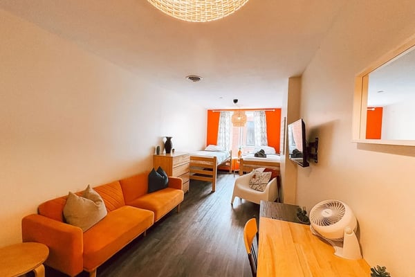 Studio apartment with orange sofa, wooden furniture, two beds with orange headboards, and woven ceiling light fixture