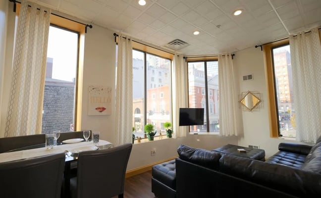 Open-plan living and dining area with two gray sofas, orange accent pillows, wooden dining table, and sunset artwork on walls